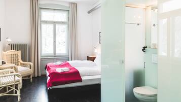 Habitación de hotel con cama, una silla, ventana con cortinas, radiador y baño con inodoro y ducha.