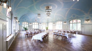 Un amplio salón de banquetes con techos altos, diseños ornamentales en el techo y múltiples candelabros. Mesas dispuestas para comer, con sillas alrededor.
