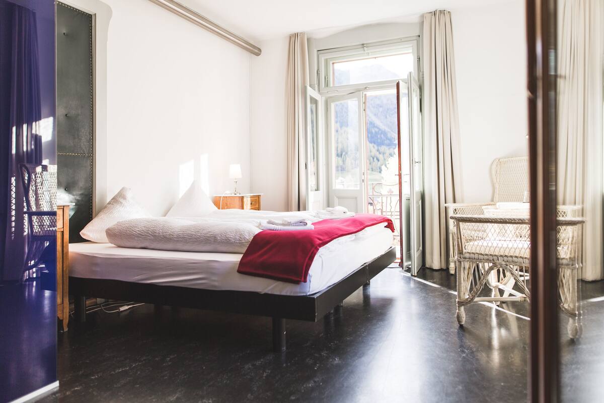 A hotel room with a bed, a nightstand, a lamp, a chair, and a view of the outdoors.
