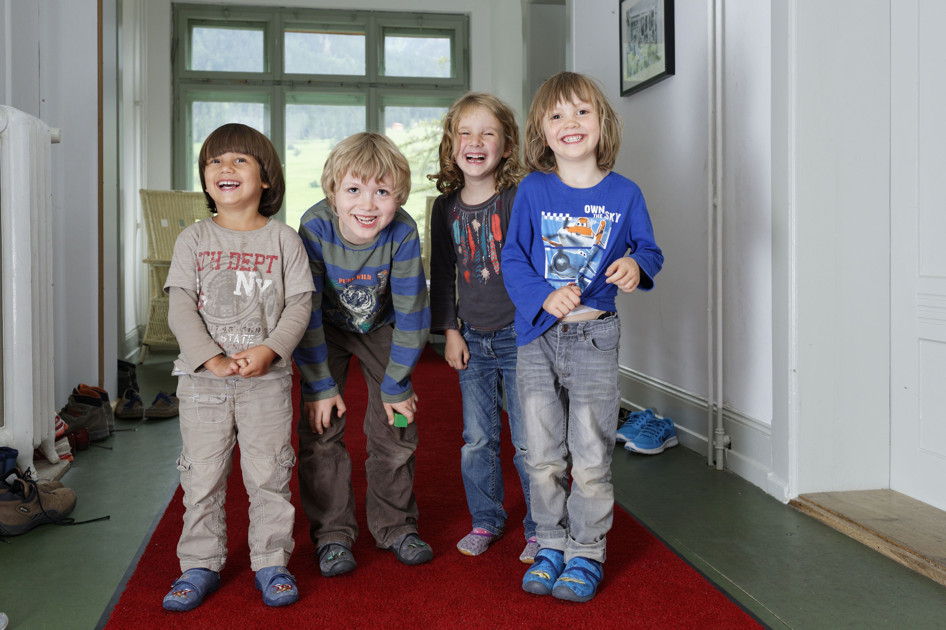 Quatre enfants se tiennent sur un tapis rouge dans une pièce avec une porte blanche et des fenêtres.