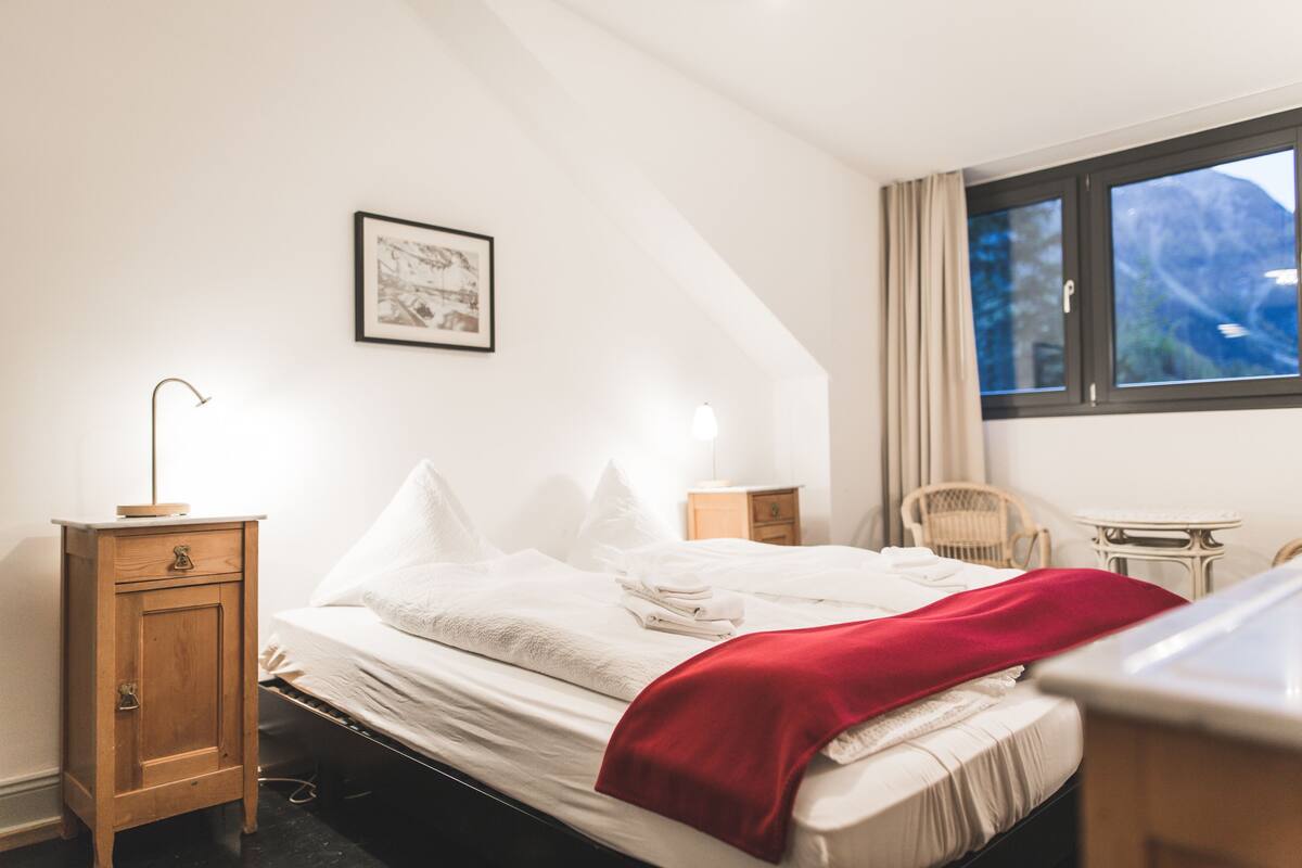 A neatly made bed with white linens and a red blanket, a wooden nightstand with a lamp, a framed picture on the wall, and a window with a view of mountains.