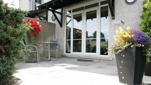 An outdoor patio with a table and chairs, a potted plant, and a building entrance with a nameplate. - Windermere Suites (Windermere)