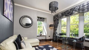 A living room with a white sofa, a round mirror, a dining table with chairs, and a view of greenery through the windows. - Windermere Suites (Windermere)