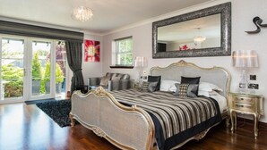 A bedroom with a large bed, a chandelier, a mirror, and a view of greenery outside. - Windermere Suites (Windermere)