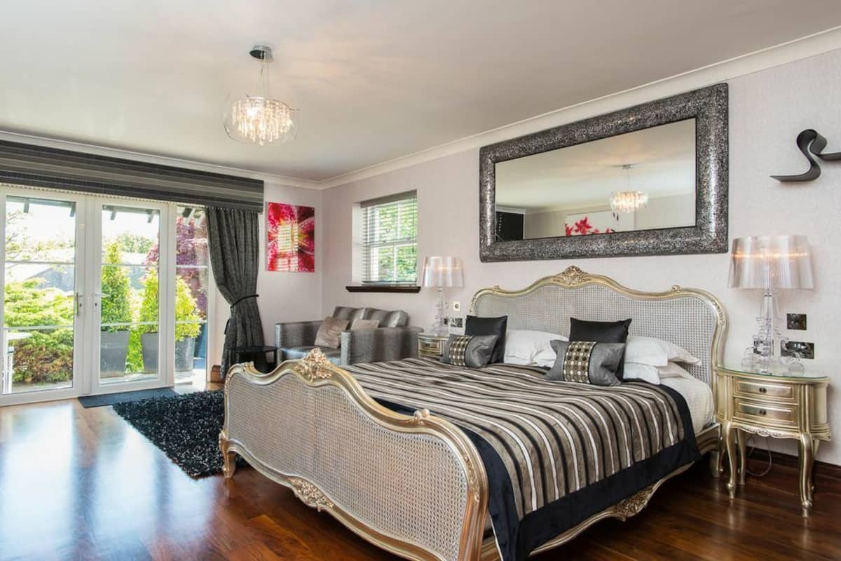 A bedroom with a large bed, a chandelier, a mirror, and a view of greenery outside.
