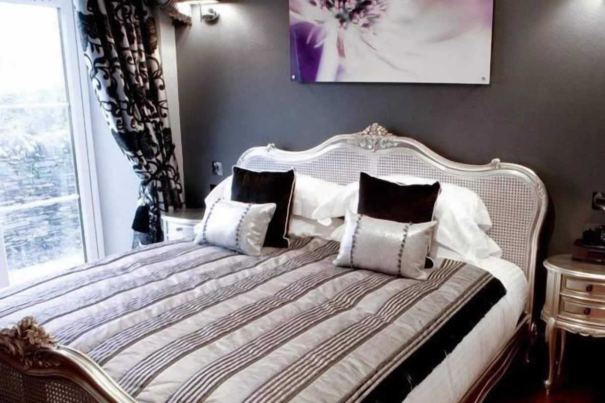 A bedroom with a large bed, a nightstand, a wall-mounted lamp, and a window with curtains.