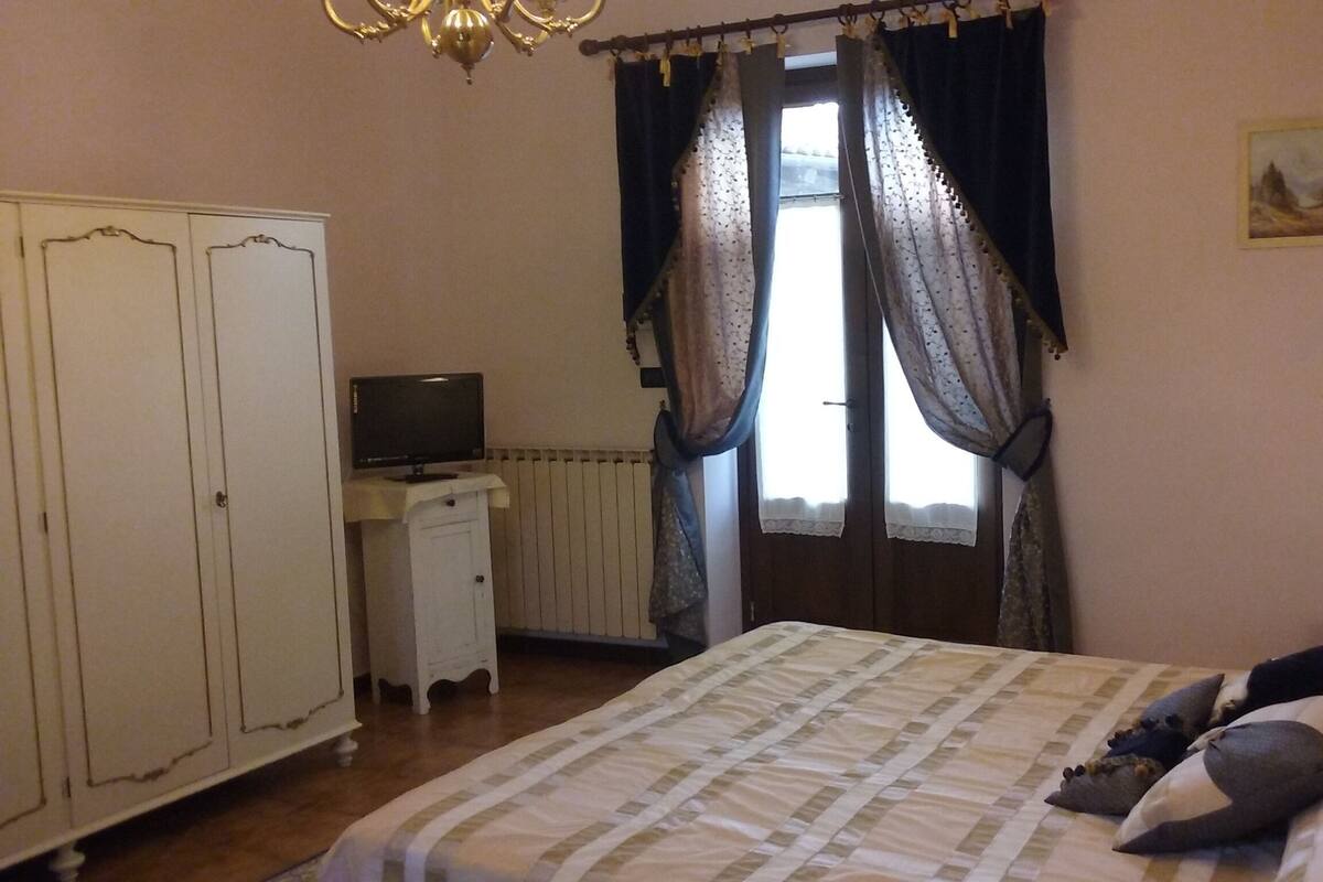 A bedroom with a bed, a television on a small table, a wardrobe, and a window with curtains.