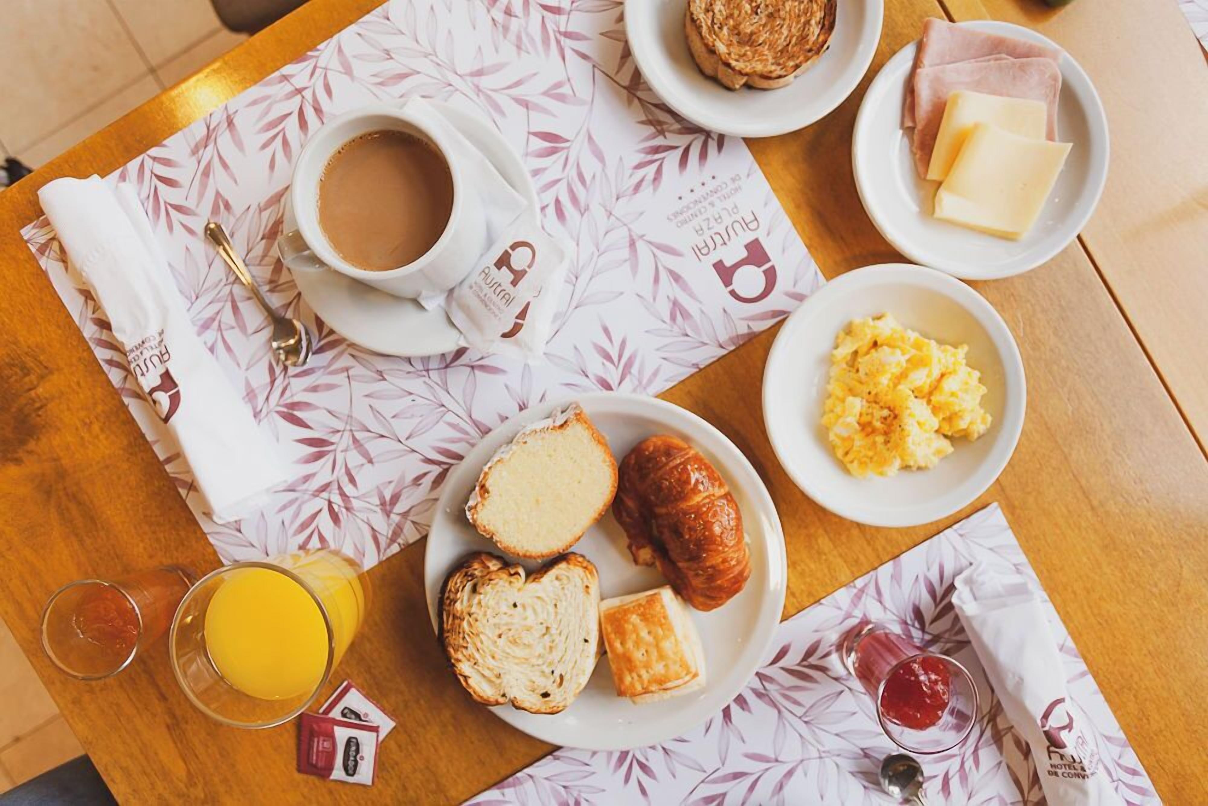 A breakfast setup with coffee, croissants, toast, scrambled eggs, ham, cheese, and juice on a table with a patterned tablecloth.