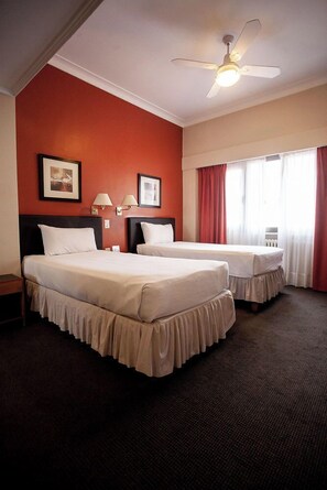 A hotel room with two beds, a ceiling fan, a window with curtains, and framed pictures on the wall.