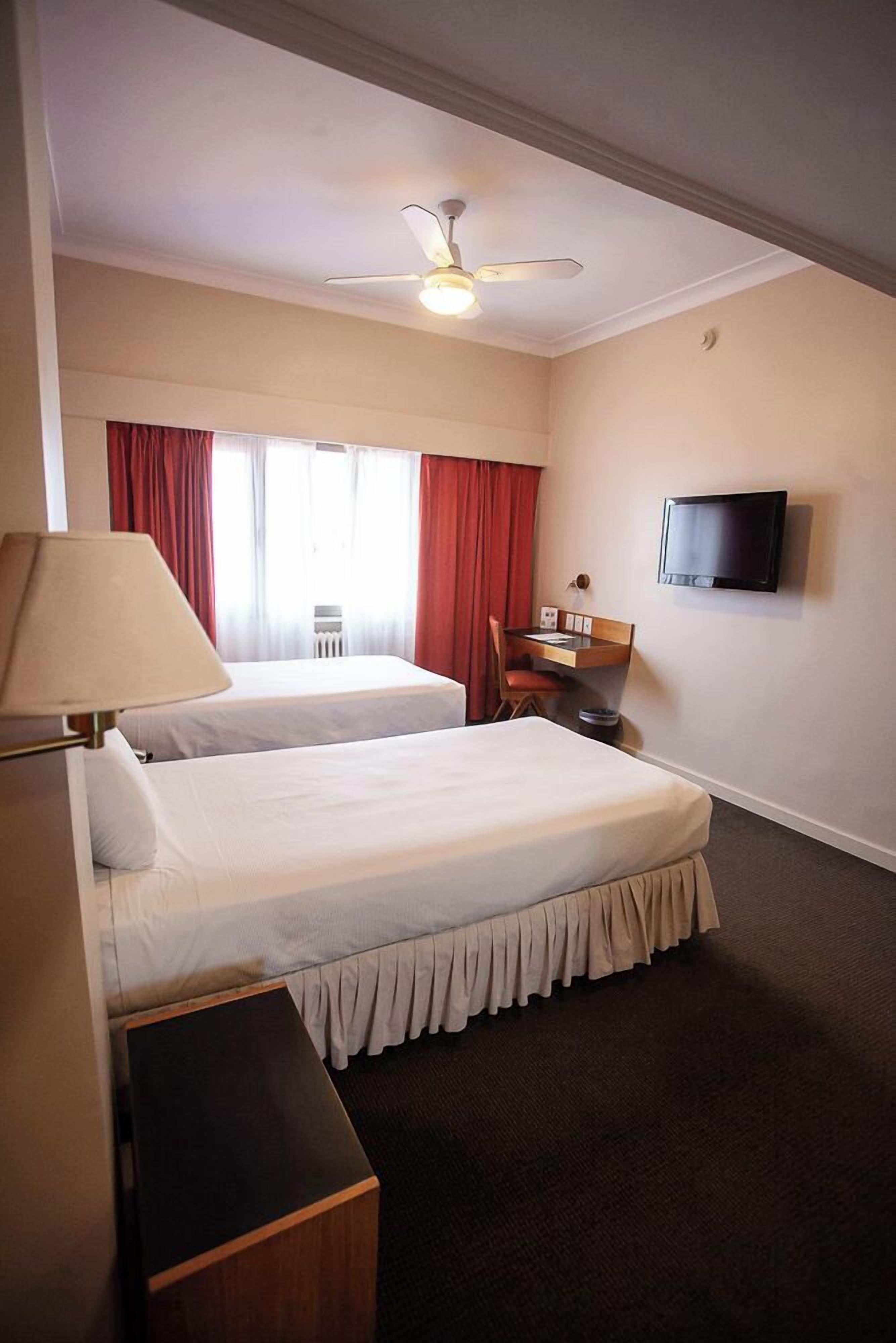 Een hotelkamer met twee bedden, een bureau, een stoel, een televisie en een plafondventilator.