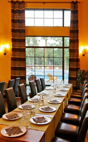 A dining area with a long table set for a meal, large windows with curtains, and a view of an outdoor area.