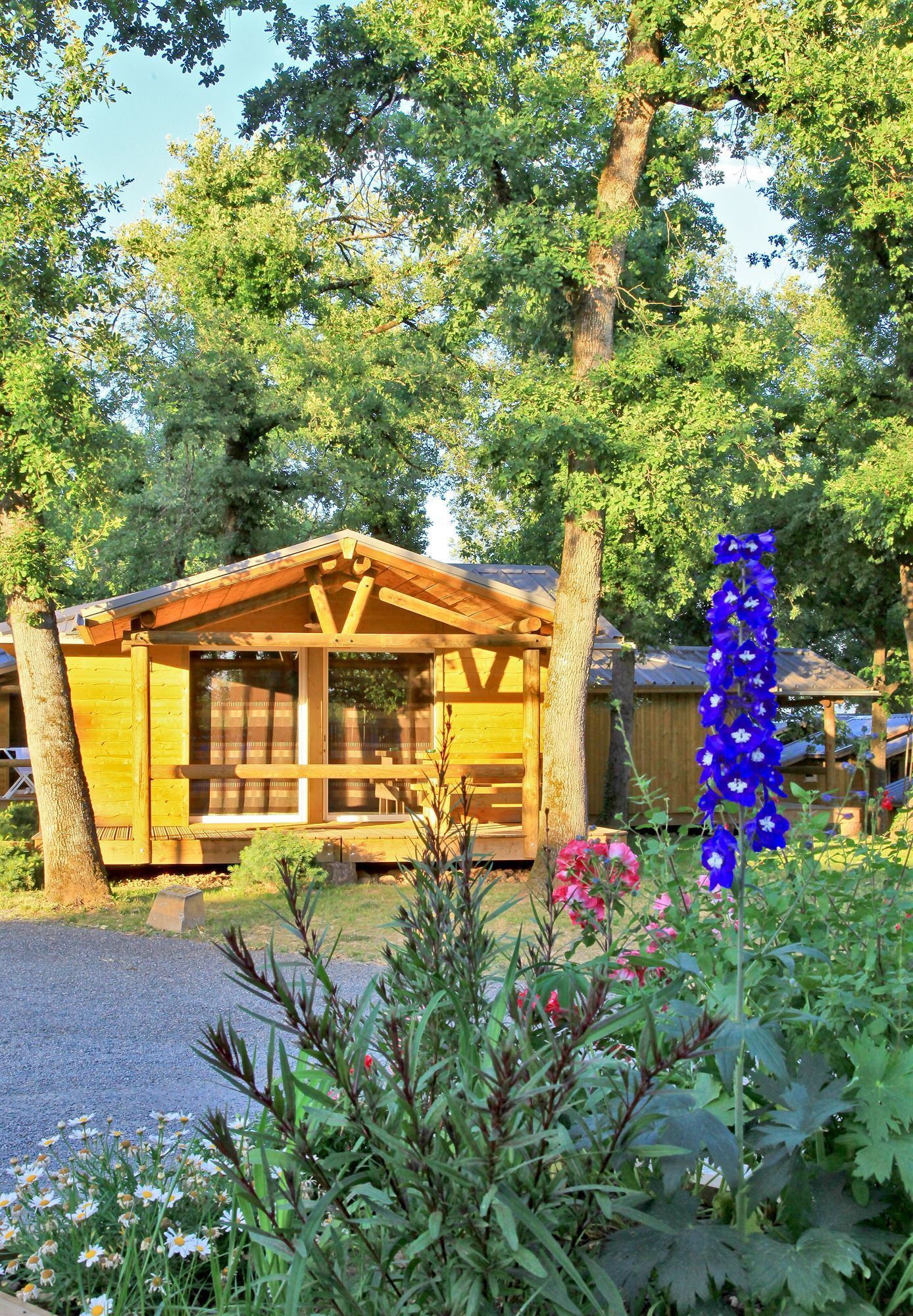 A wooden cabin with a porch, surrounded by trees and a garden with flowers.