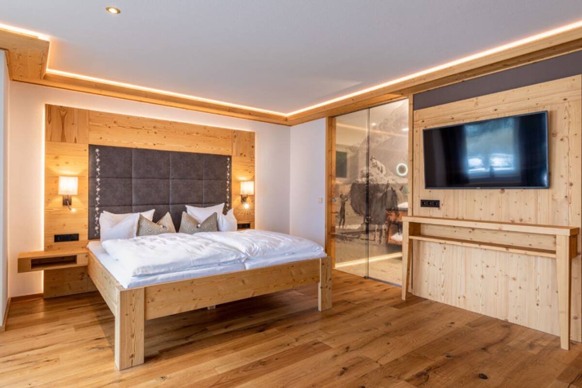 Un dormitorio moderno con una cama de madera, un televisor montado en una pared de madera y una puerta de cristal que da al baño.