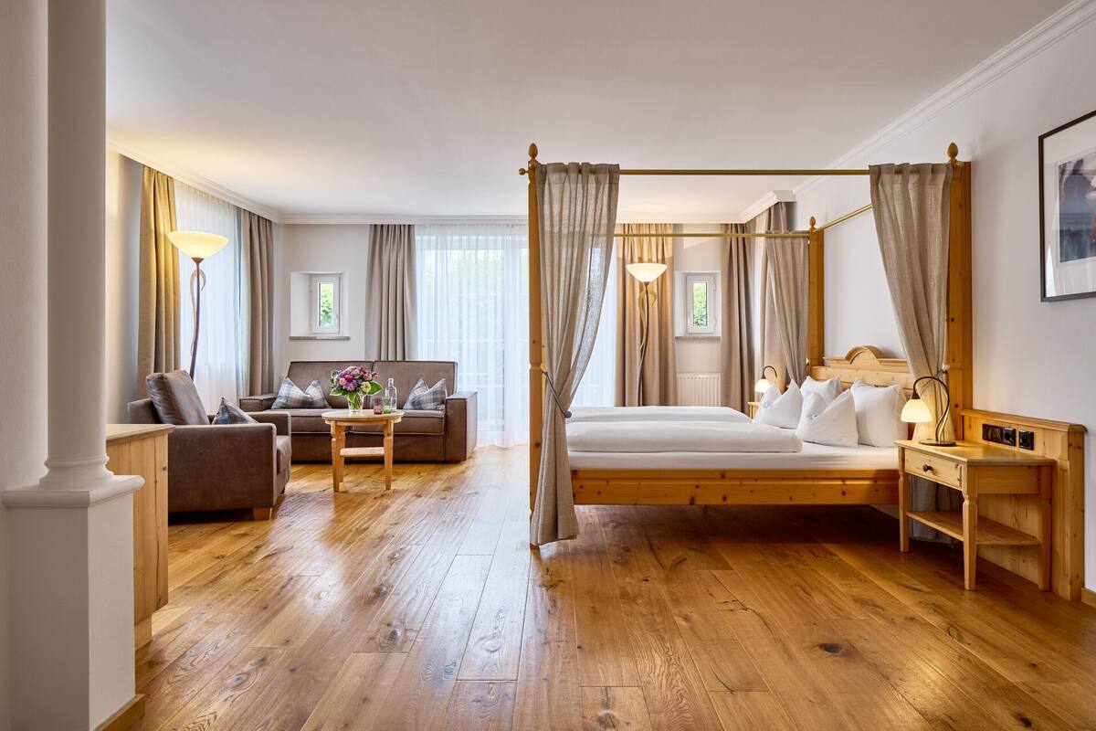 Un dormitorio espacioso con cama con dosel, una zona de estar con sofá y una mesa, suelo de madera y ventanas grandes con cortinas.