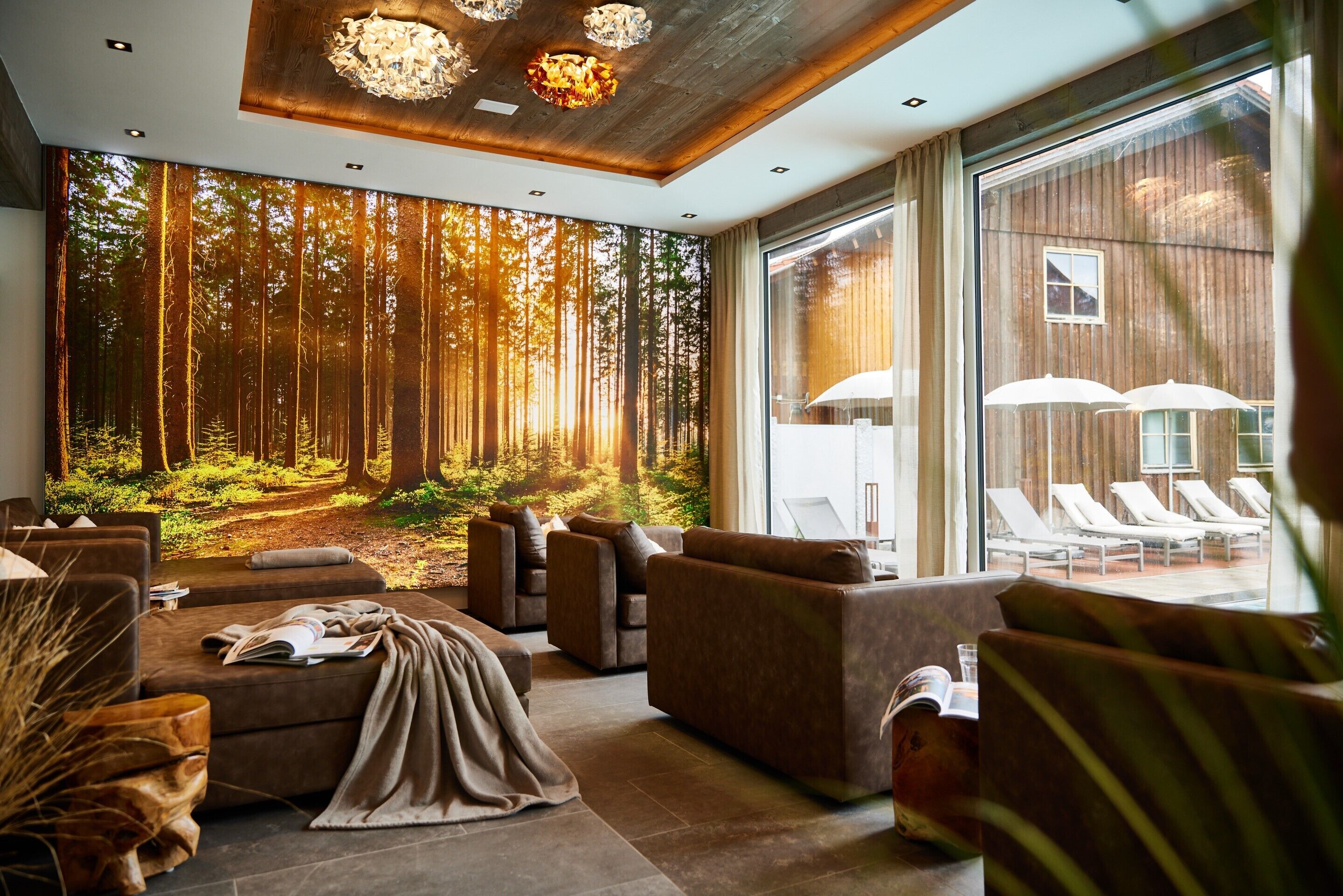Un área de lounge moderna con un mural temático de bosque, asientos cómodos y grandes ventanales con vistas a una terraza exterior.