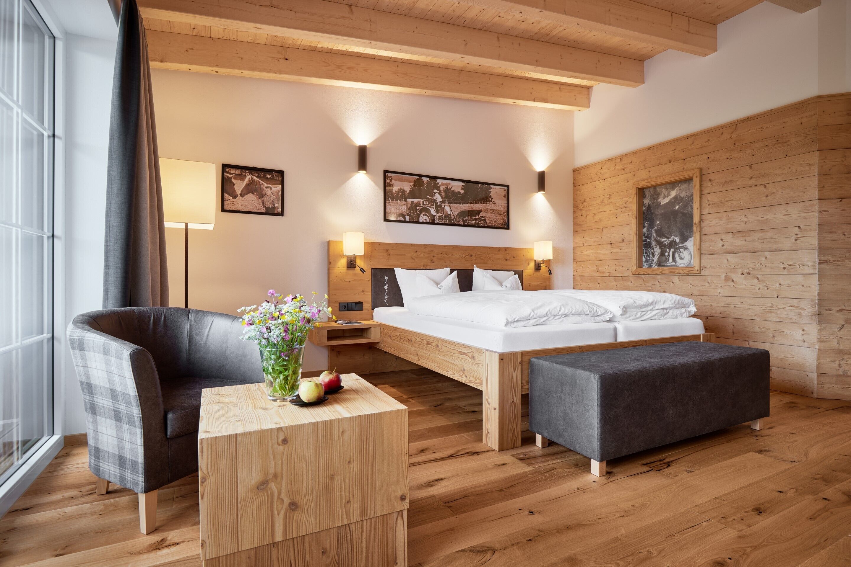 A modern bedroom with a wooden ceiling, a bed with white linens, a grey armchair, a wooden side table with a vase and fruit, and two wall-mounted lamps.