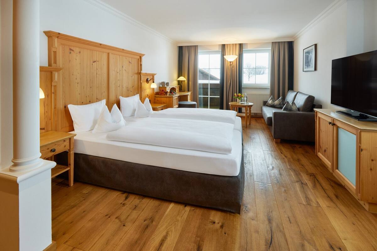 Habitación de hotel con cama doble, sofá, televisión, suelo de madera y ventana con cortinas.