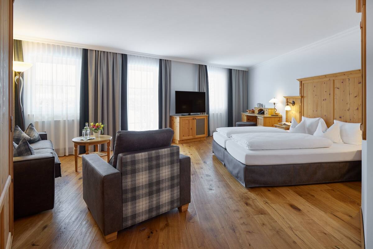 Habitación de hotel con sofá, una silla, cama, televisor, una mesita con lámpara y un mueble de madera.
