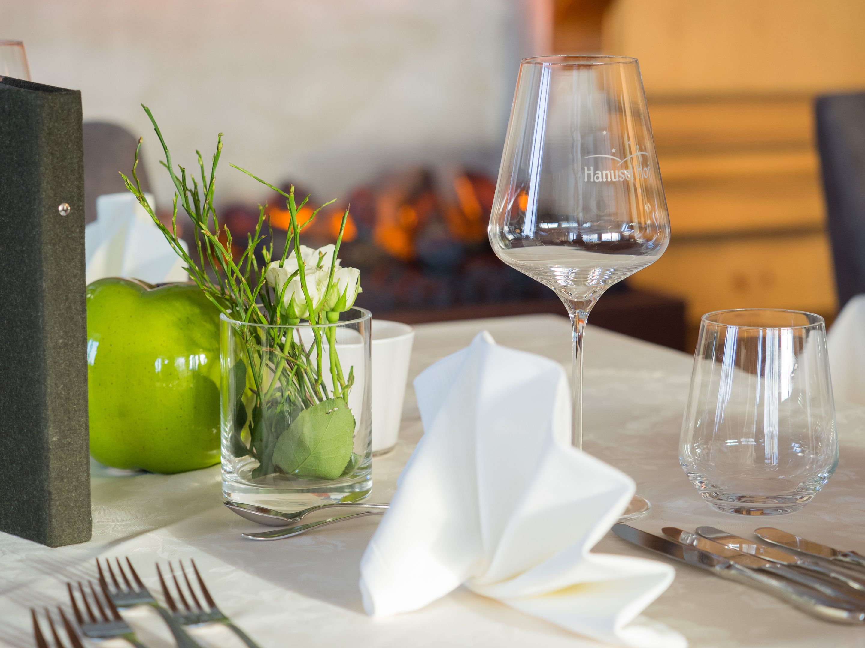 Una tavola imbandita con una mela verde, un vaso di vetro con steli e fiori bianchi, un bicchiere da vino e posate.