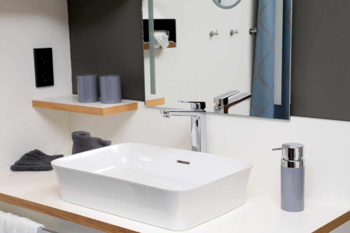 Une salle de bain moderne avec un lavabo blanc, un robinet chromé et un miroir au-dessus. On peut voir deux serviettes sur le comptoir et un distributeur de savon à droite.