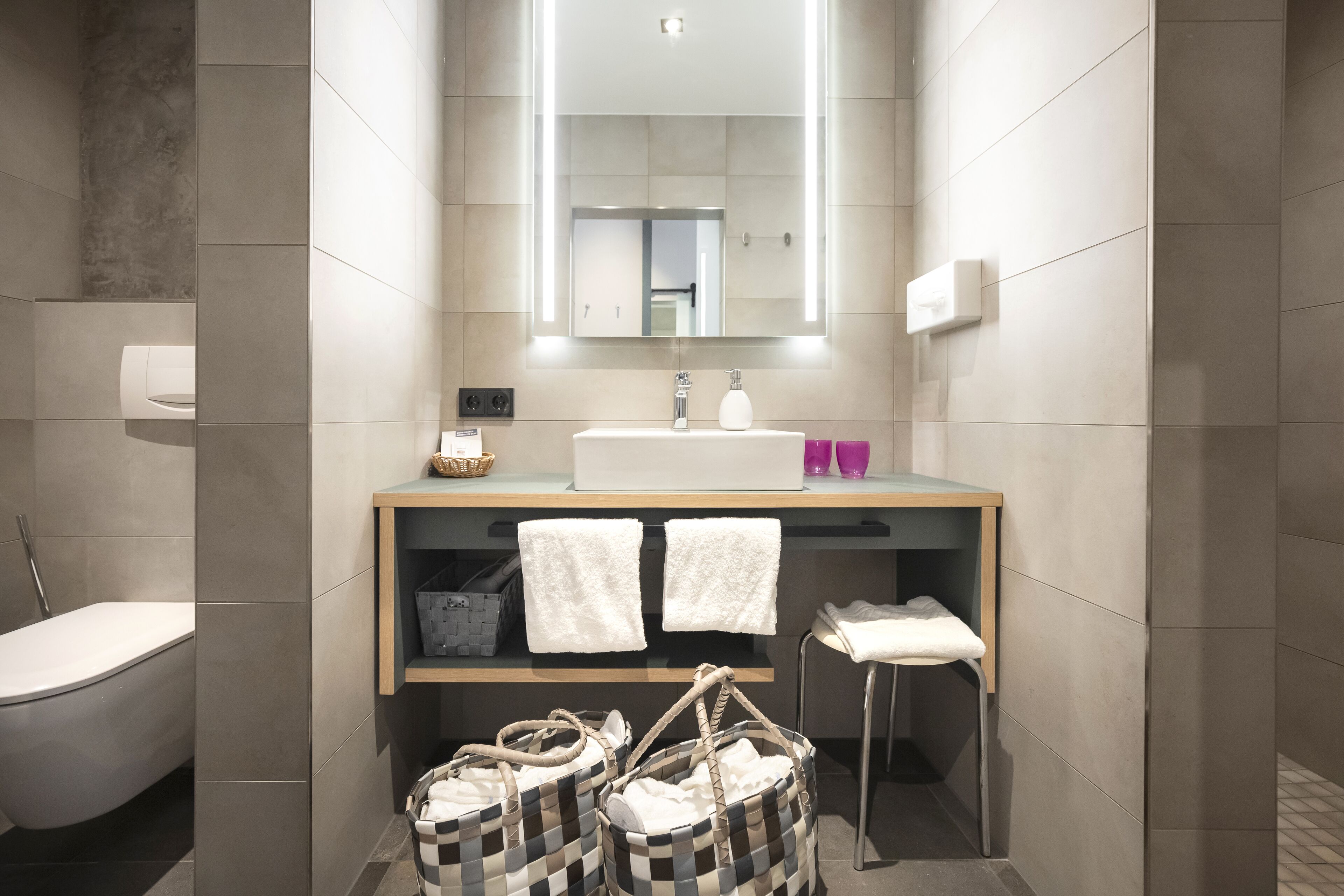 Ein modernes Badezimmer mit einem weißen Waschbecken, einem Spiegel und einem Handtuchhalter. Zwei weiße Handtücher hängen am Halter und es gibt ein kleines Holzregal mit einem Korb und einem Handtuch.