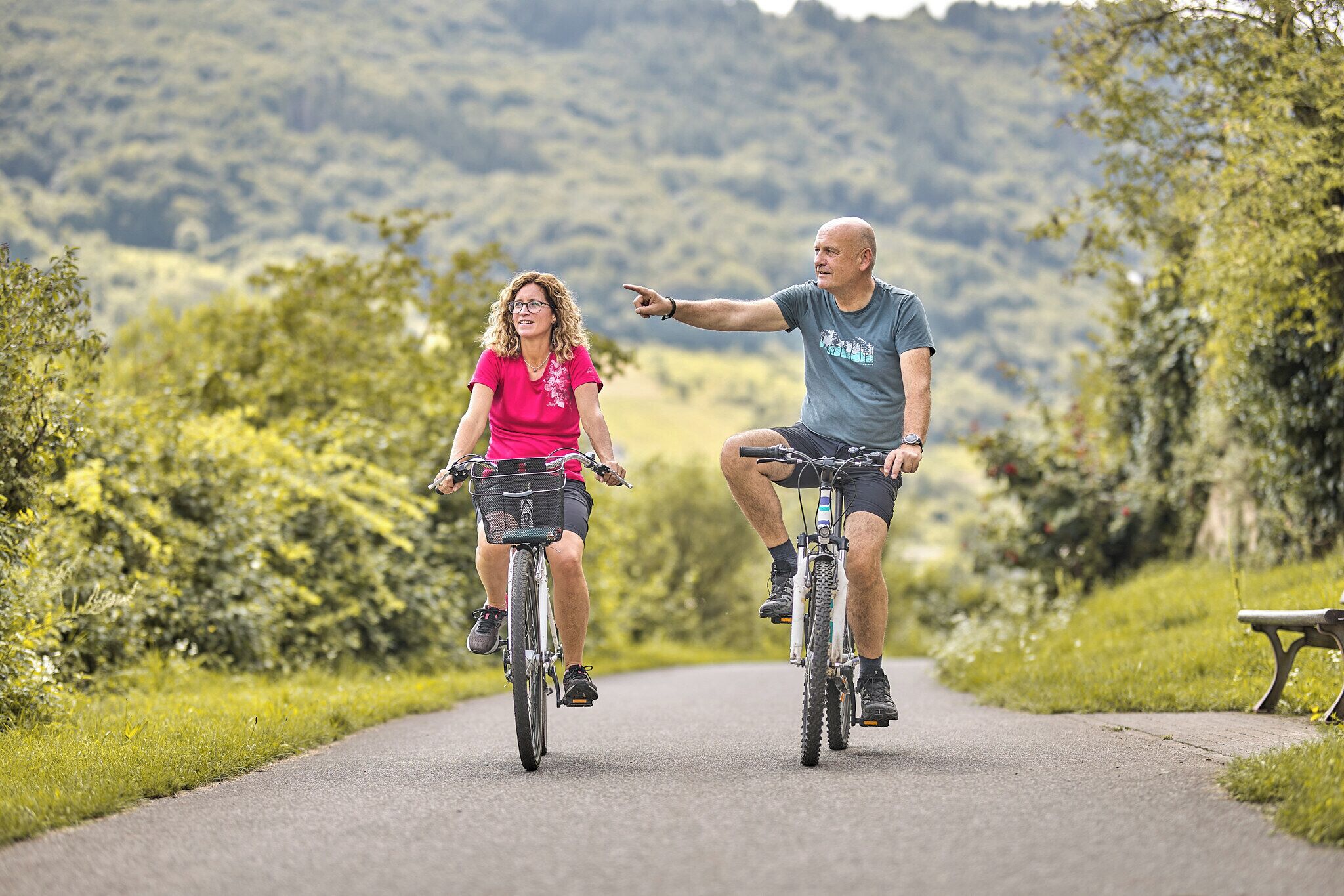 Duas pessoas de bicicleta em um caminho pavimentado, rodeadas por vegetação.