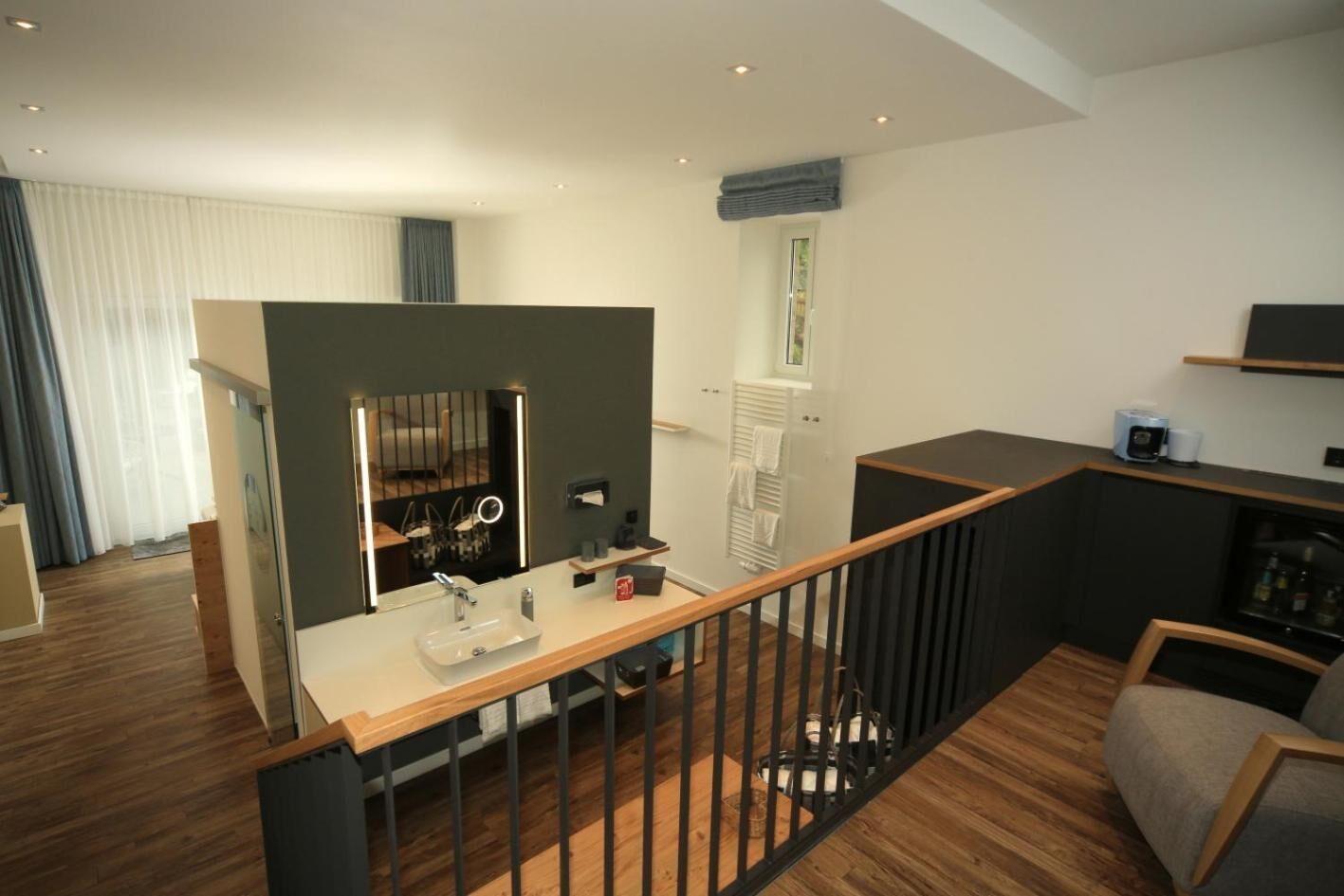 Una sala de estar moderna con piso de madera, un área de comedor con una mesa blanca y sillas negras, una cocina con cubierta negra y una escalera con barandilla de madera.