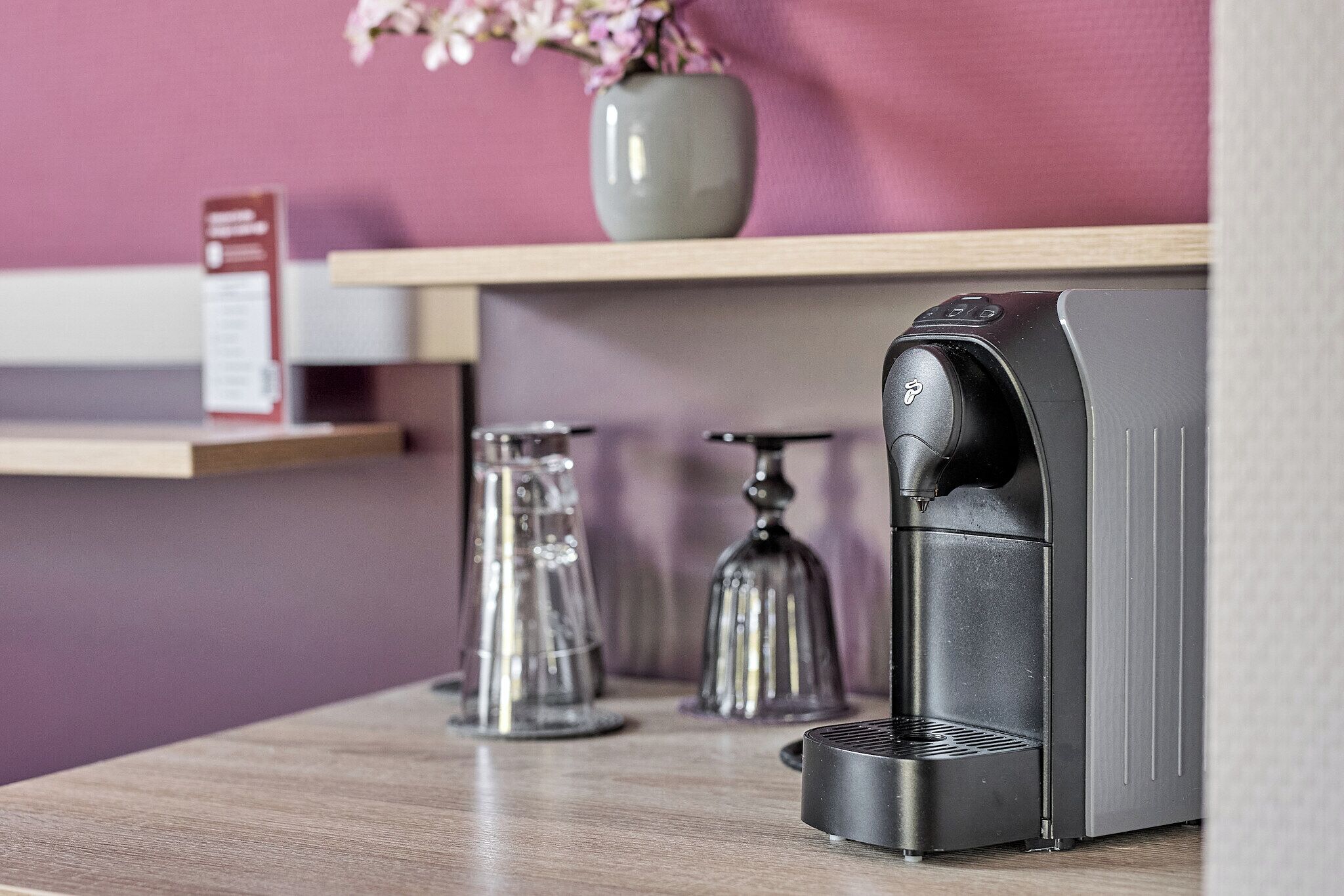 Een koffieapparaat op een houten aanrecht, met een bloevas en een bord in de achtergrond.