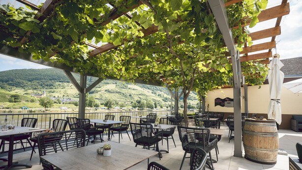Une terrasse extérieure avec des tables et des chaises, une pergola en bois et une vue sur un paysage vallonné.
