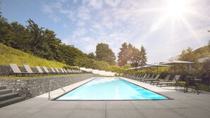 Une piscine rectangulaire bordée de pierres, entourée de chaises longues et de parasols, avec en arrière-plan un paysage de arbres et de collines.
