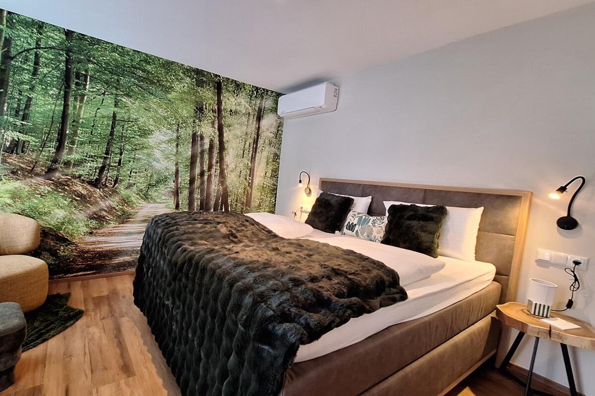 A bedroom with a forest-themed wall mural, a bed with a headboard, bedside lamps, and a small bedside table.