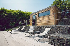 Un moderno espacio lounge al aire libre con terraza de madera y paredes de piedra, que incluye sillones y un área de descanso junto a una pared de piedra.