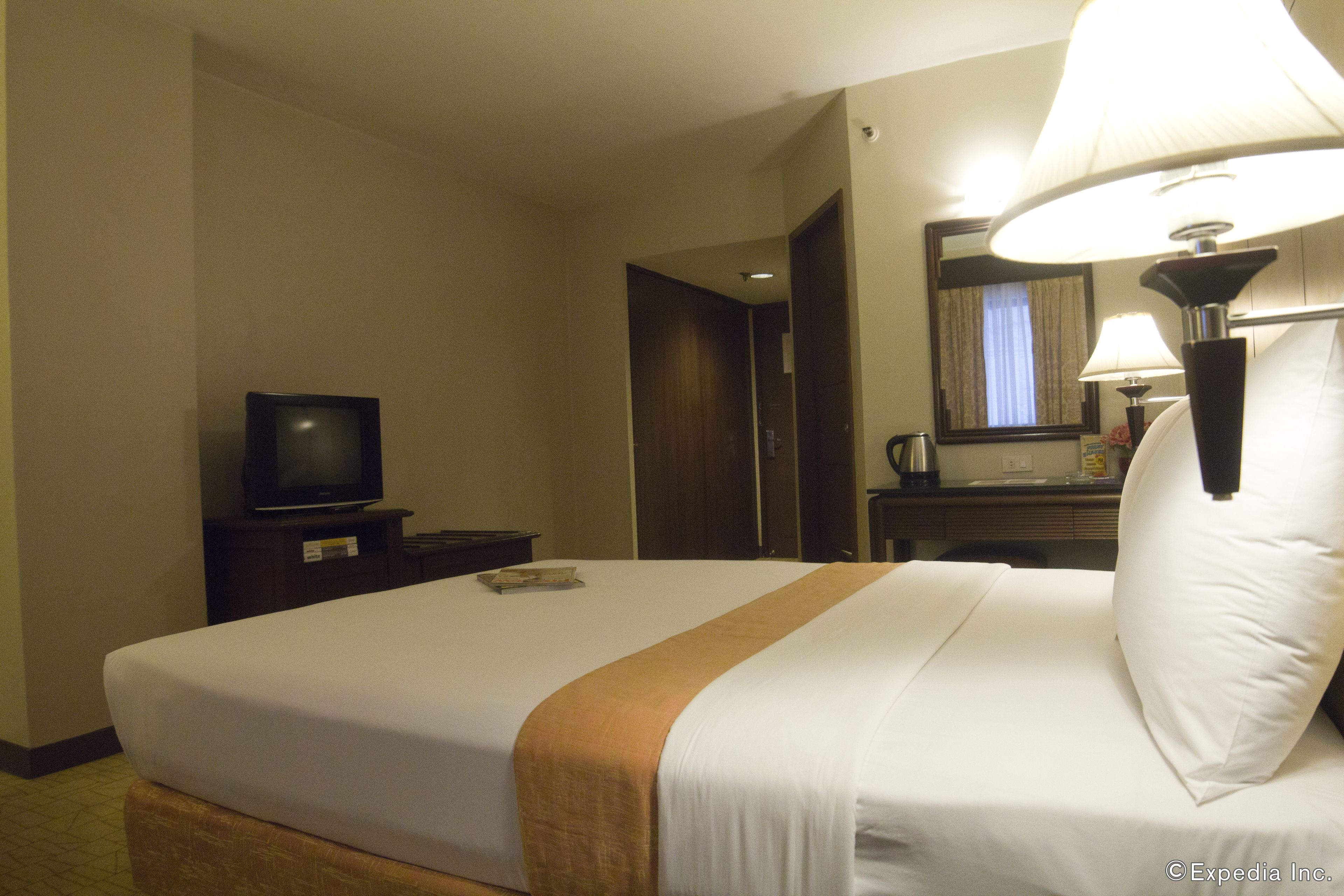 A hotel room with a bed, a television on a nightstand, a lamp, and a mirror.