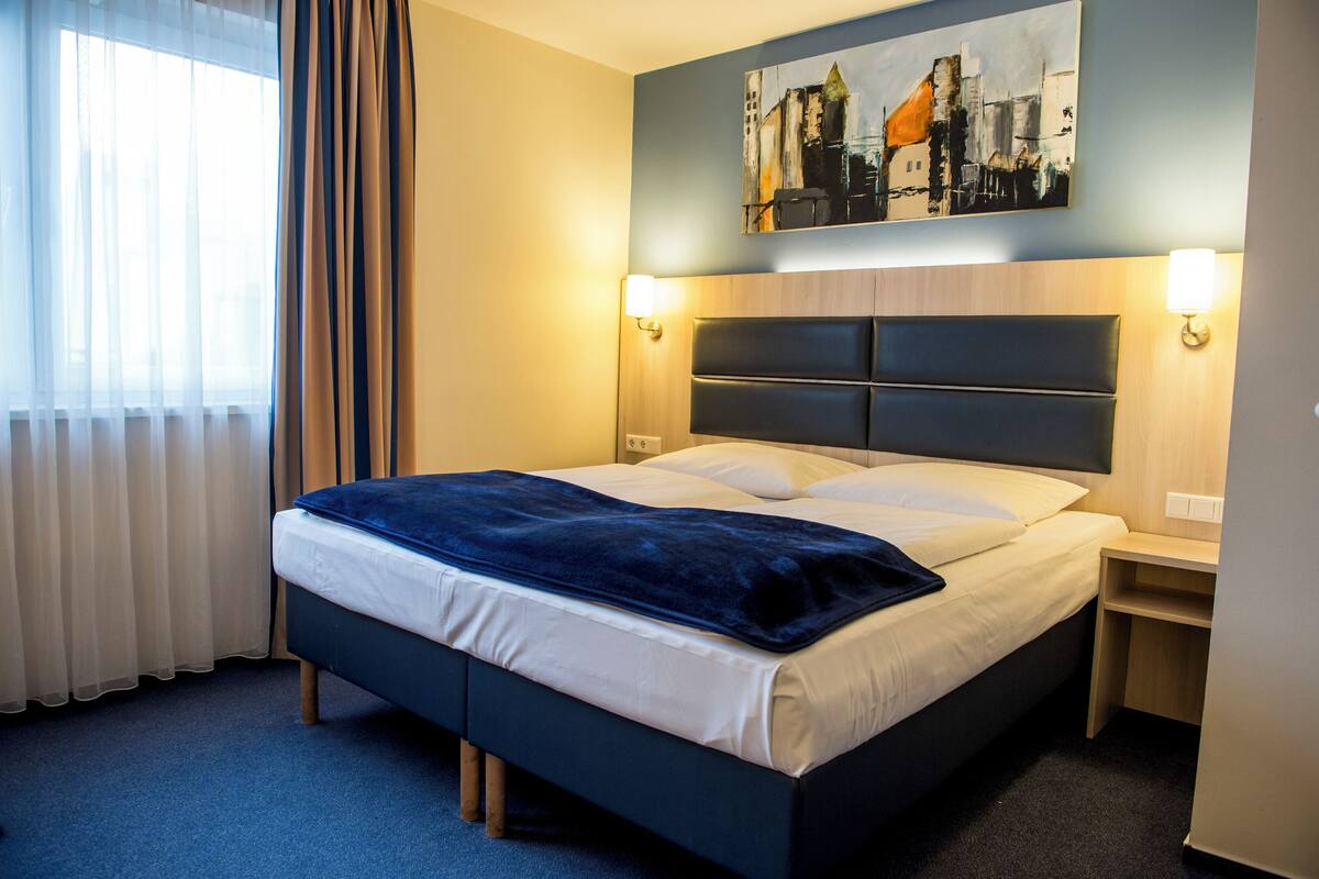 A neatly made bed with a dark headboard, a bedside table with a lamp, and a painting above the bed.