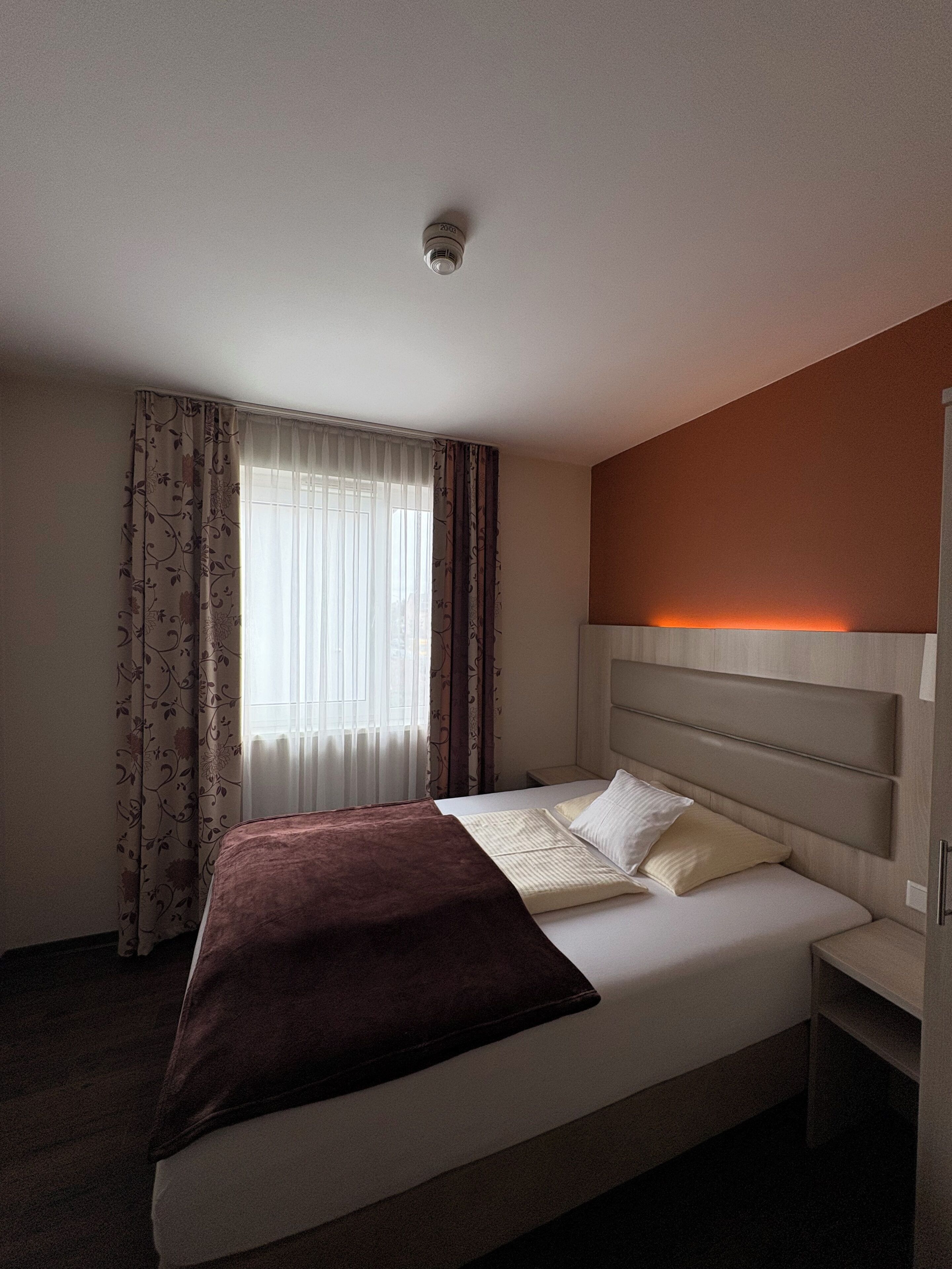 A hotel room with a bed, curtains, a headboard, and a ceiling light.