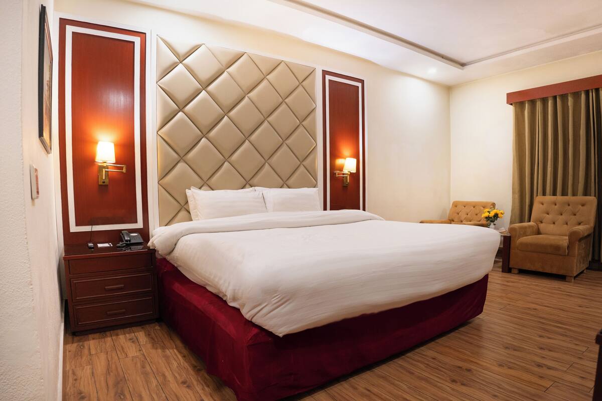 Hotelkamer met een groot bed, een gestoffeerd hoofdbord, twee bedlampen, een houten nachtkastje, een rode sprei en een bruine fauteuil, op een houten vloer.
