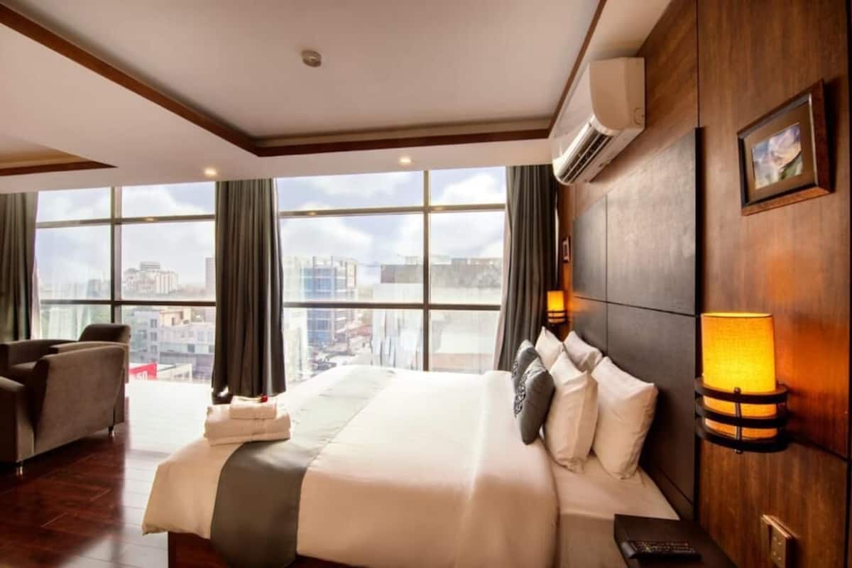 Habitación de hotel con una cama grande, un sofá, un escritorio y vistas a la ciudad a través de amplias ventanas.