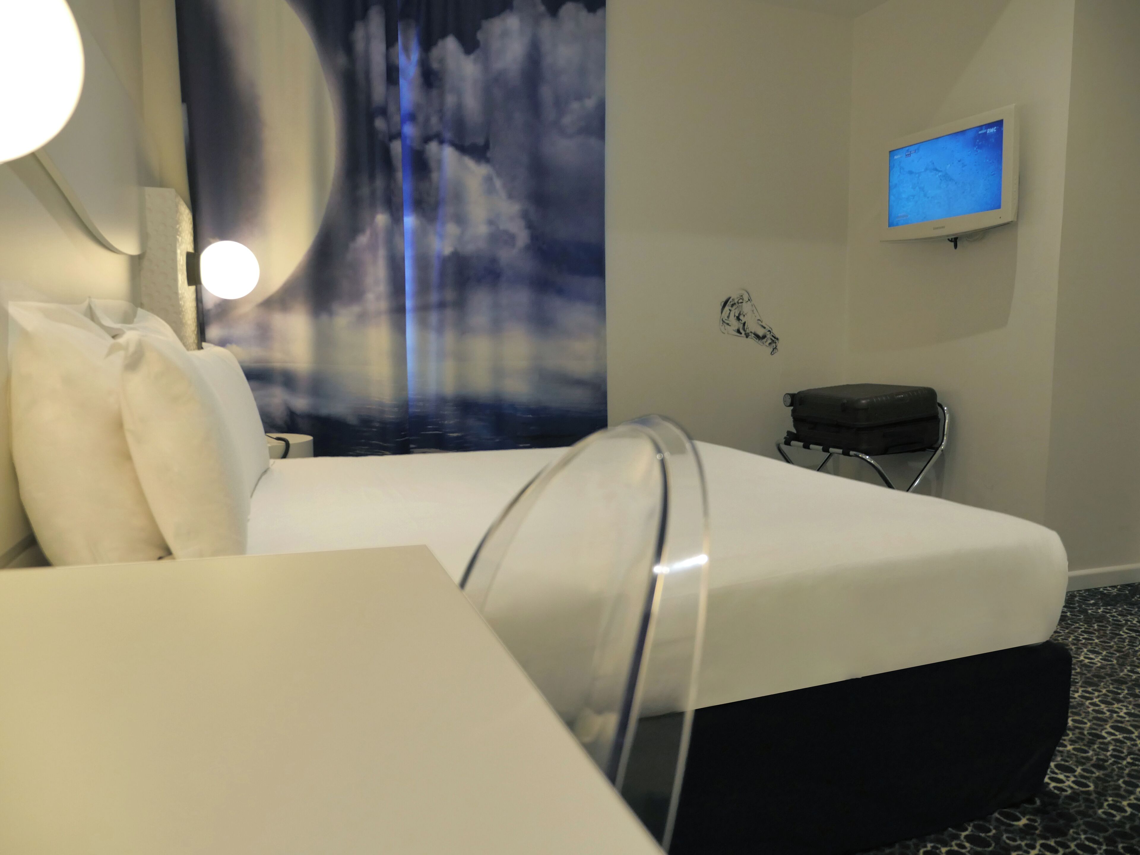 Hotellihuone, jossa on sänky, seinälle asennettu taulutelevisio, pieni pöytä ja tuoli.