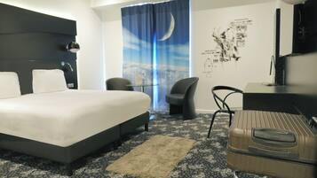 A hotel room with a large bed, a desk, a chair, a suitcase, a small table with a bottle, and a wall with a poster.