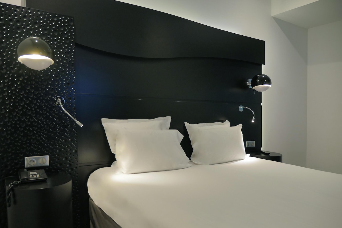 A modern hotel room with a large bed, two white pillows, a black headboard, a round wall-mounted light, a bedside table with a phone, and a small nightstand.