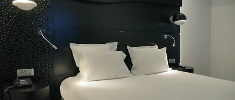 A modern hotel room with a large bed, two white pillows, a black headboard, a round wall-mounted light, a bedside table with a phone, and a small nightstand.