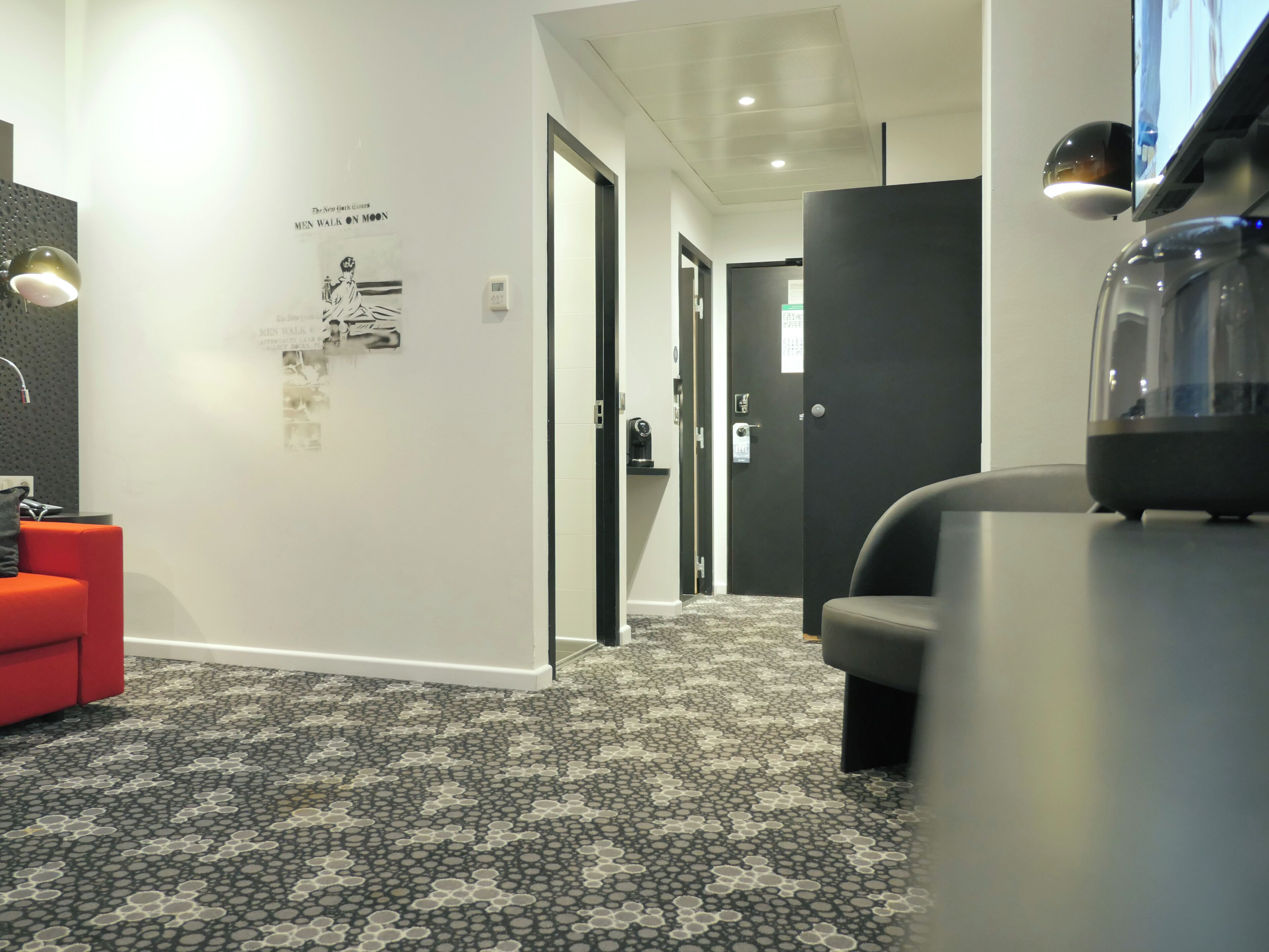A modern hotel lobby with a patterned carpet, a red seating area, a reception desk, and a television screen.