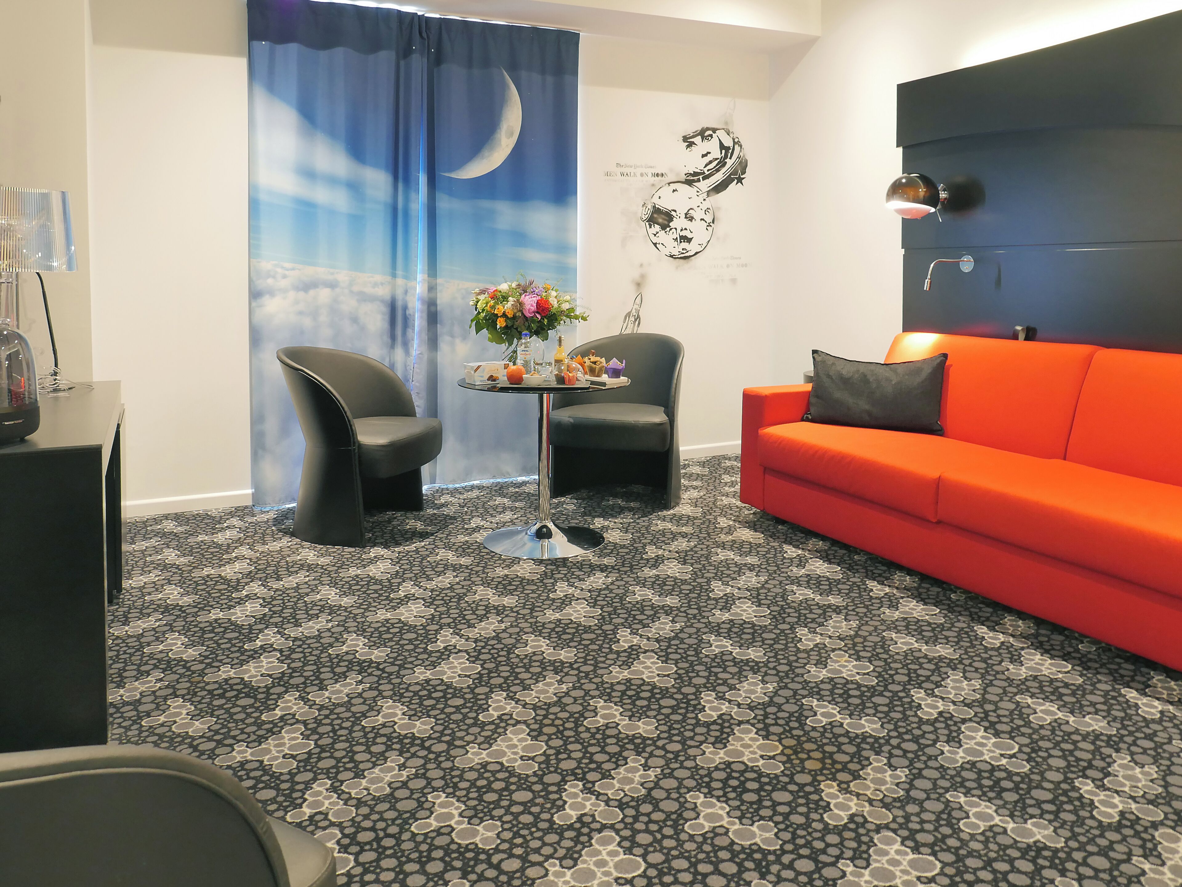 A modern living room with a black and white patterned carpet, a red sofa, two black chairs, a small round table with a flower vase, and a wall with a moon and stars mural.