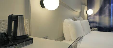 A hotel room with a bed, pillows, a bedside table with a kettle, and wall-mounted lights.