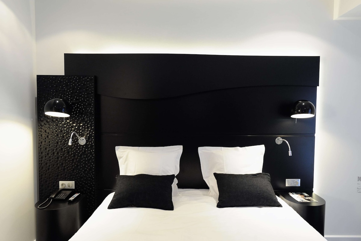 A modern hotel room with a large bed, two bedside tables, a black headboard, and a textured wall panel.