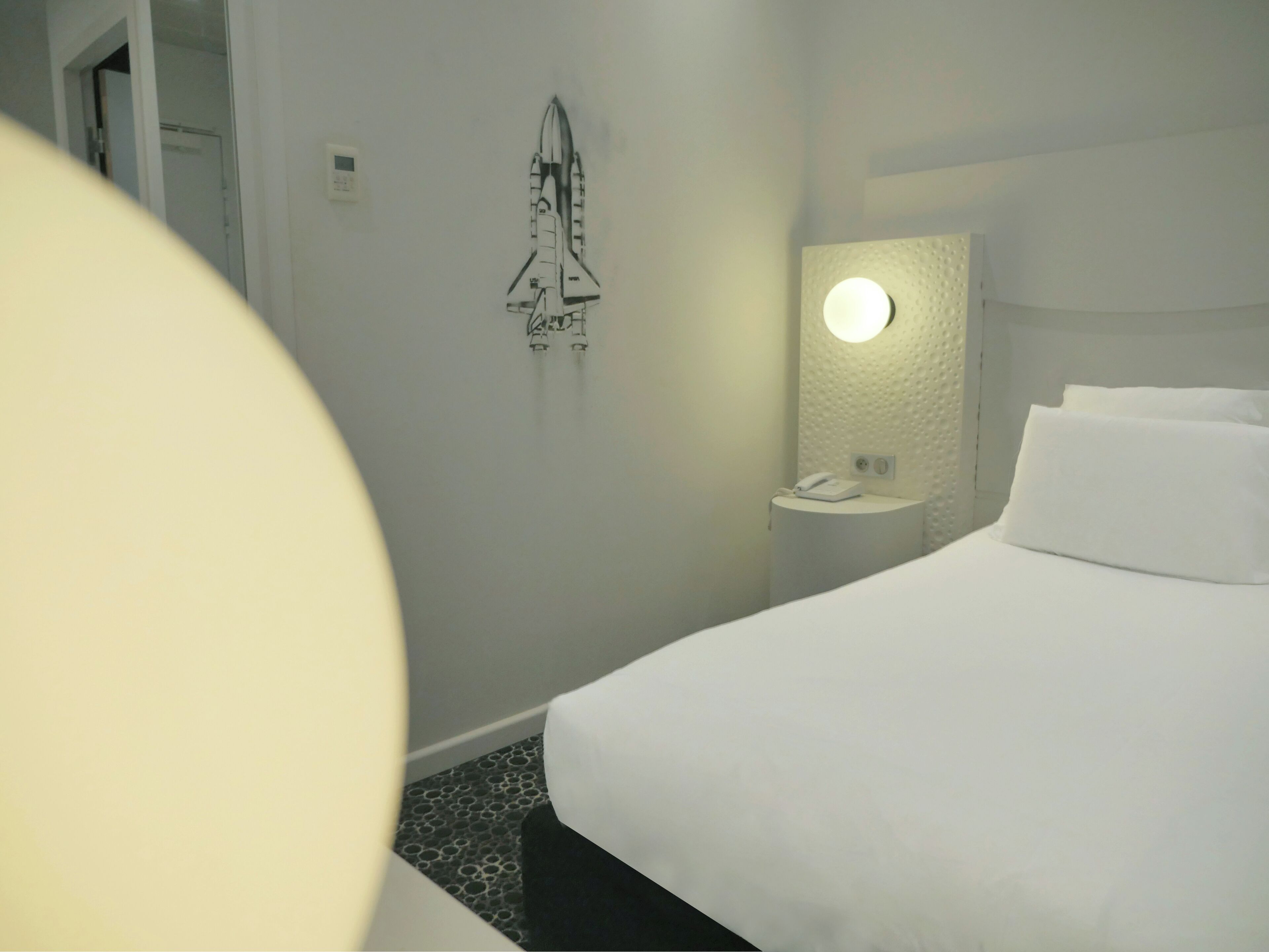 Habitación de hotel con cama, mesita de noche con teléfono, luz empotrada en la pared y una decoración con transbordador espacial.