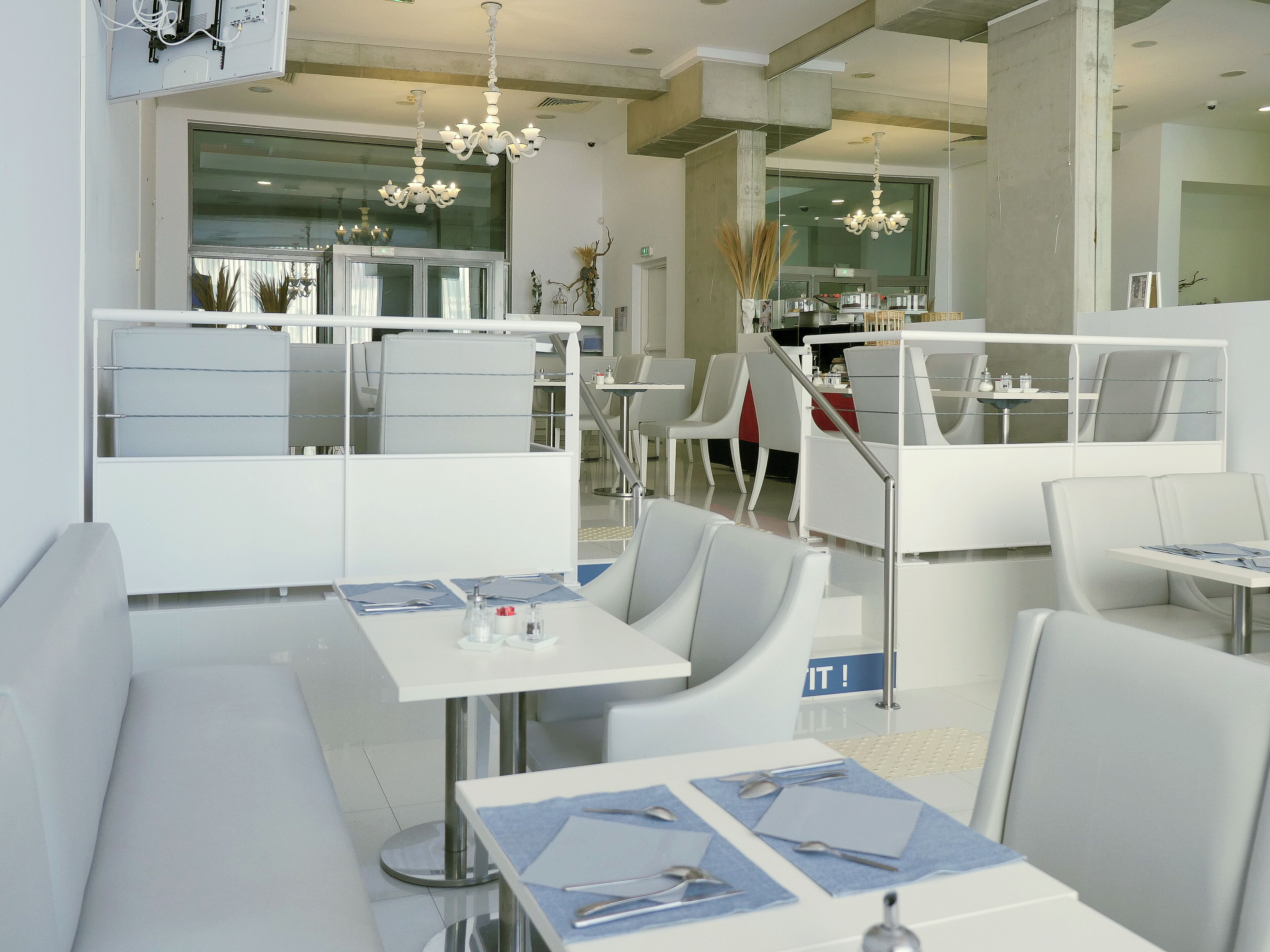 A modern restaurant with white furniture, a chandelier, and a glass partition separating the dining area from the kitchen.