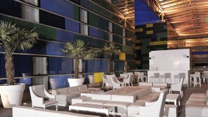 Un espacio moderno al aire libre con mobiliario blanco, plantas en macetas grandes y un edificio contemporáneo con ventanales y techo de madera.