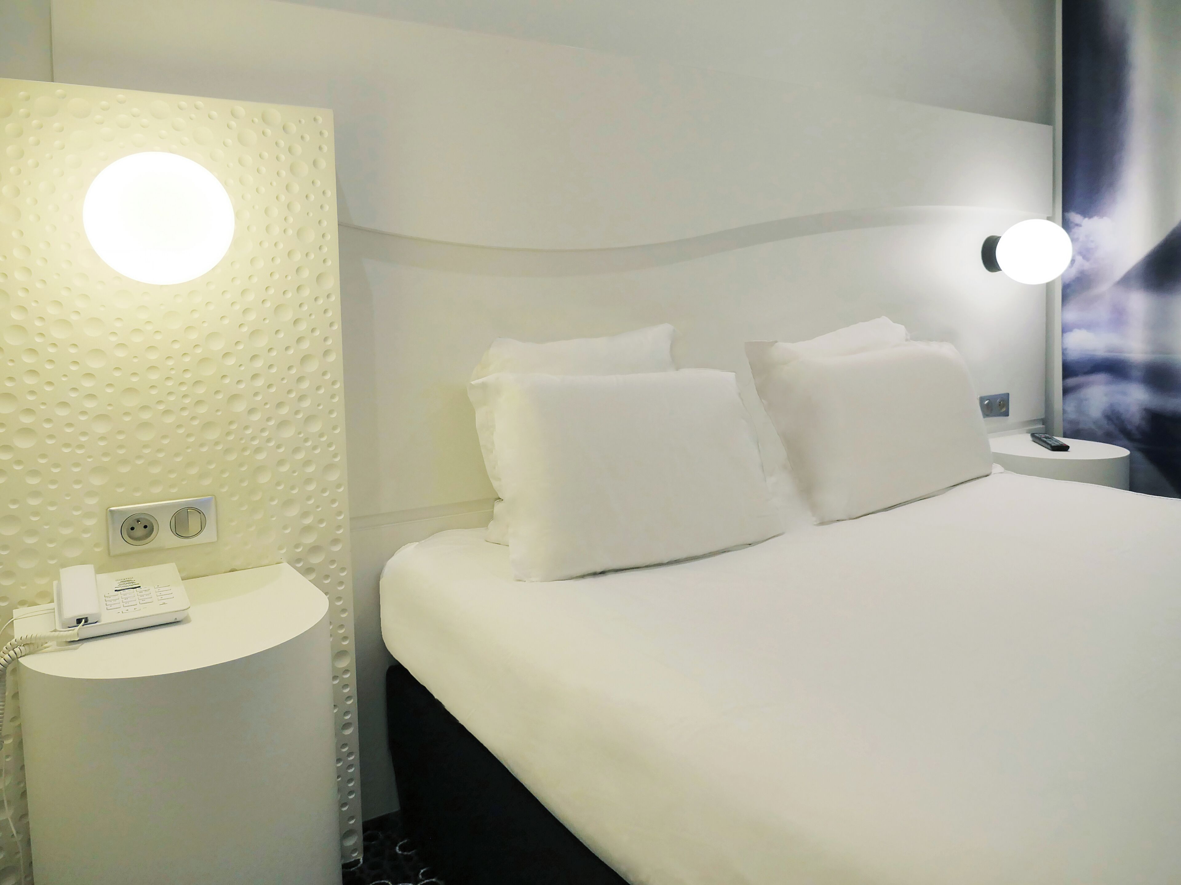 A hotel room with a bed, a bedside table with a phone, and a wall-mounted light fixture.