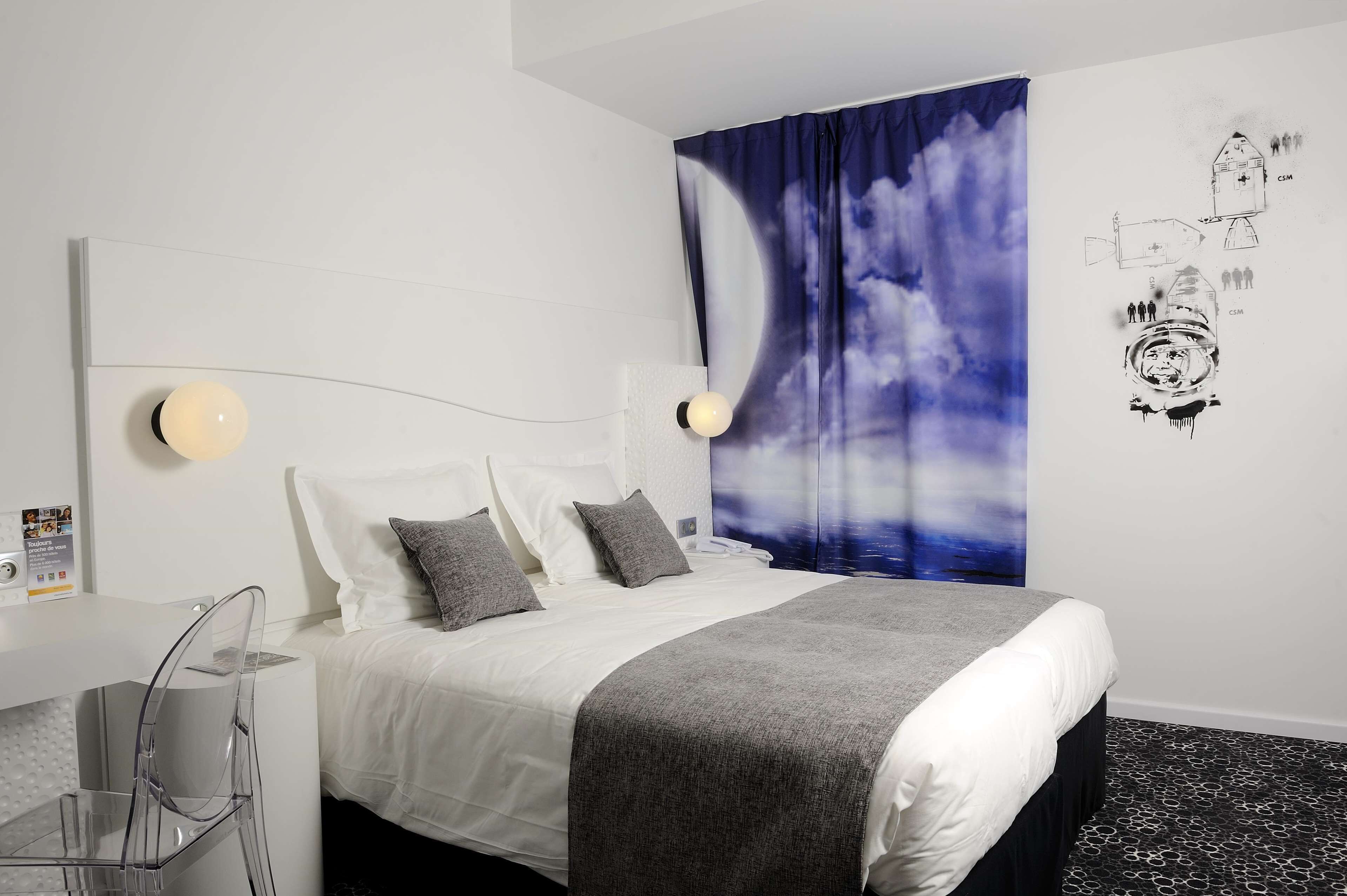 A modern hotel room with a neatly made bed, a desk with a chair, and a large wall tapestry depicting a space theme.